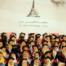 Women students wearing graduation caps and gowns sit in front of Al Aqsa University banner