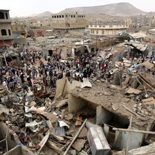 A crowd of people stands in the ruins of a bombed-out marketplace