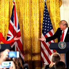 Theresa May and Donald Trump, standing at podiums during White House press conference, smile and reach their arms towards one another