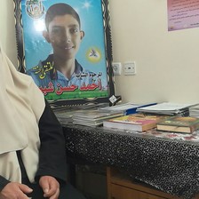 Woman with covered face sits next to table and poster of her smiling teenage son