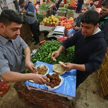 Two men stand at table making falafel sandwiches in middle of produce shop