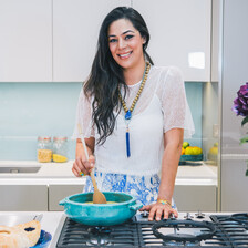 Smiling young woman seen waist-up standing behind kitchen counter while stirring pot on a stovetop