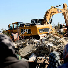 Photo shows the backs of women's heads as they watch a Caterpillar machine being used to raze a home