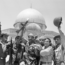 Soldiers seen from waist up raise rifles and helmets above their head while standing in front of Dome of the Rock