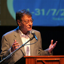 Seen from waist up, Mahmoud Darwish gestures with his hands as he stands behind a podium on a stage