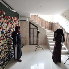 Woman wearing niqab takes a photo of a smiling woman standing in front of a wall decorated with paper flowers