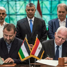 Two men sitting at a desk sign documents as three men standing behind them look on