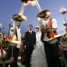 A bride and groom walk through a procession of men in traditional dress carrying burning torches