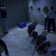 Six men wearing hoods in a dark room are put in various stress positions while a seventh man wearing a guard's uniform stands next to one of them