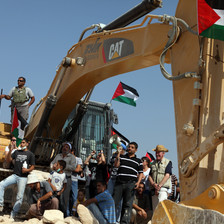 A multigenerational group of people, some carrying Palestine flags, sit around an earth-moving machine with Caterpillar's logo on it and an Israeli soldier standing on top of it