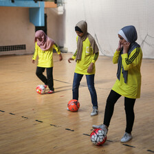 Three girls manipulate footballs with their feet in a gymnasium