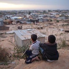 Two boys sit on a sand embankment overlooking a village of corrugated metal shelters