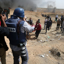 Photographer wearing flak jacket marked as PRESS and wearing a blue helmet is seen from behind while taking photos of demonstrators