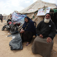 Elderly man and woman sit in front of tent adorned with banner stating We are here to return to our lands