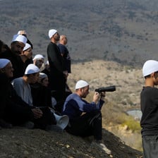 A group of men and boys wearing traditional white caps stand and sit on hillside looking over boundary fence, some holding binoculars