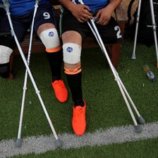 Photo shows legs and crutches of men in football uniform sitting on a bench at a football pitch