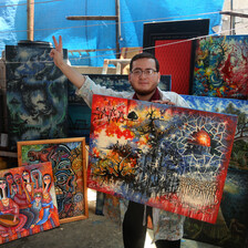 Young man holding large painted canvas gives the victory hand gesture as he stands in a room surrounded by his paintings