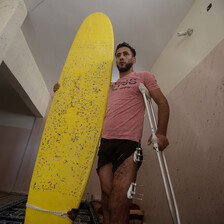 Man wearing t-shirt and shorts holds a surfboard with one arm and supports himself with crutches using his other arm