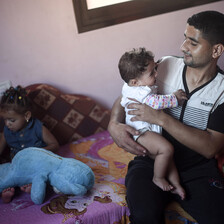 Muhammad al-Najjar smiles and looks at his infant daughter who he is holding while his toddler daughter sits on couch behind him