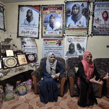 Two women sit on chairs in room with walls covered with posters of Razan al-Najjar in medic's uniform