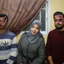Alaa Abu Mustafa stands between two of her "zombies." One her right, a man has his face apaprently split open, with bones protruding. On her left, a man's eye is made out to be dangling down his cheek. 