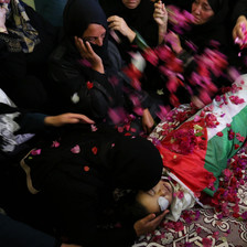 Women lean over and throw flower petals on body of boy shrouded in Palestinian flag