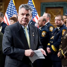 Peter King, a member of Congress for New York, stands beside US flags and soldiers in uniform.