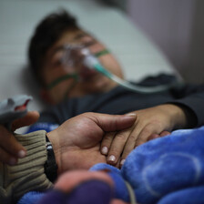 Young patient on a hospital bed, with a mask on his face and his hand being held by another person.