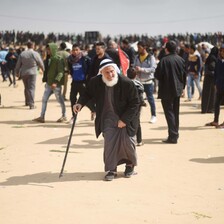 Man wearing white headdress walks with aid of stick surrounding by various people. 