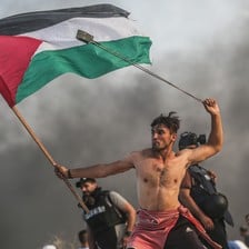 Bare-chested man wields slingshot over his head while holding Palestine flag