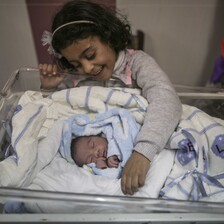 A little girl smiles at a newborn baby wrapped in a blanket.