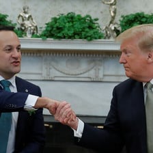 Man wearing tie shakes hands with another man wearing green tie; a white marble fireplace decorated with shamrock and statues can be seen behind them.