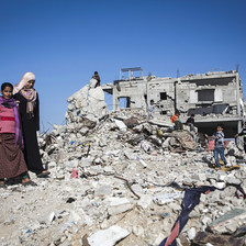 Women and children stand on the remains of a bombed-out building