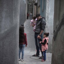 A man carrying a young girl stands flanked by two other children in a narrow grey alley.