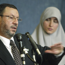 Man speaks before two microphones, woman stands next to him. 