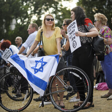 A woman wearing a yellow top holds a bicycle with a blue and white flag draped over it, while talking to another woman holding a placard that reads "Labour: For the Many, Not the Jew." 