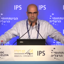 A bald man in a shirt stands at a conference podium speaking into a mic