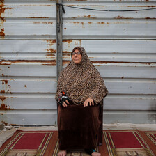 Woman sits in front of a wall, with a dark red rug beneath her feet. 