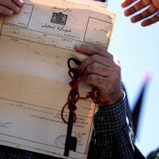 A man's hands with a key and chain wrapped around them holding up a document in Arabic