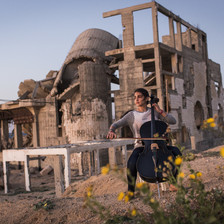 Woman plays cello in front of the rubble of a destroyed building