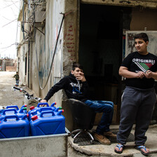 Two boys are next to several large blue plastic water canisters.