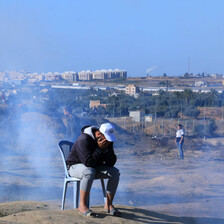 A man sits outside on a plastic chair as smoke billows behind him.