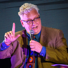 A man in a suit holding a microphone points up one finger