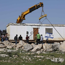 A crane hovers over a prefabricated building, surrounded by large rocks 