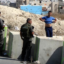 Two soldiers behind concrete blocks look at a young man who has lifted his t-shirt to show his stomach