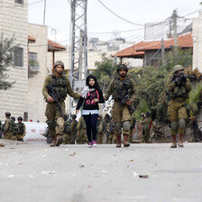 Two armed soldiers walk with a young girl between them while a third points a rifle somewhere in the distance