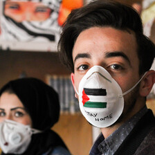 A man and a woman wear painted face masks one bearing the Palestinian flag and  words stay at home