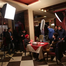 Film crew members stand around male and female actors sitting on opposite sides of a restaurant table