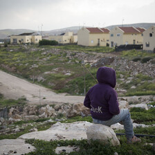 Youth sits with back turned to camera in front of Israeli settlement