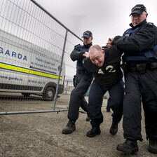 Police officers wearing baseball hats grab hold of a man by his arms 